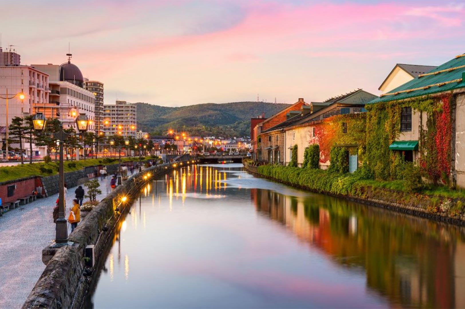 Scenic evening view of the Otaru Canal in Hokkaido, Japan, featuring historic ivy-covered stone warehouses and glowing street lamps at sunset.