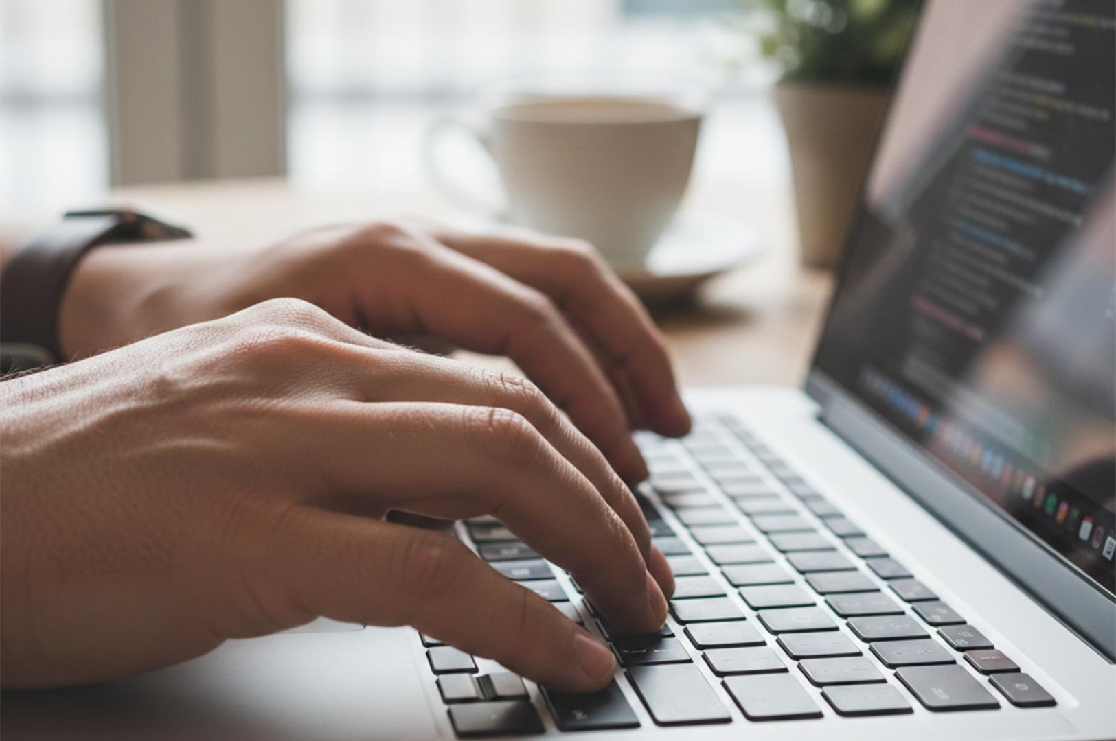 Close-up of a person's hands typing on a laptop keyboard with lines of code visible on the screen and a coffee cup in the background.