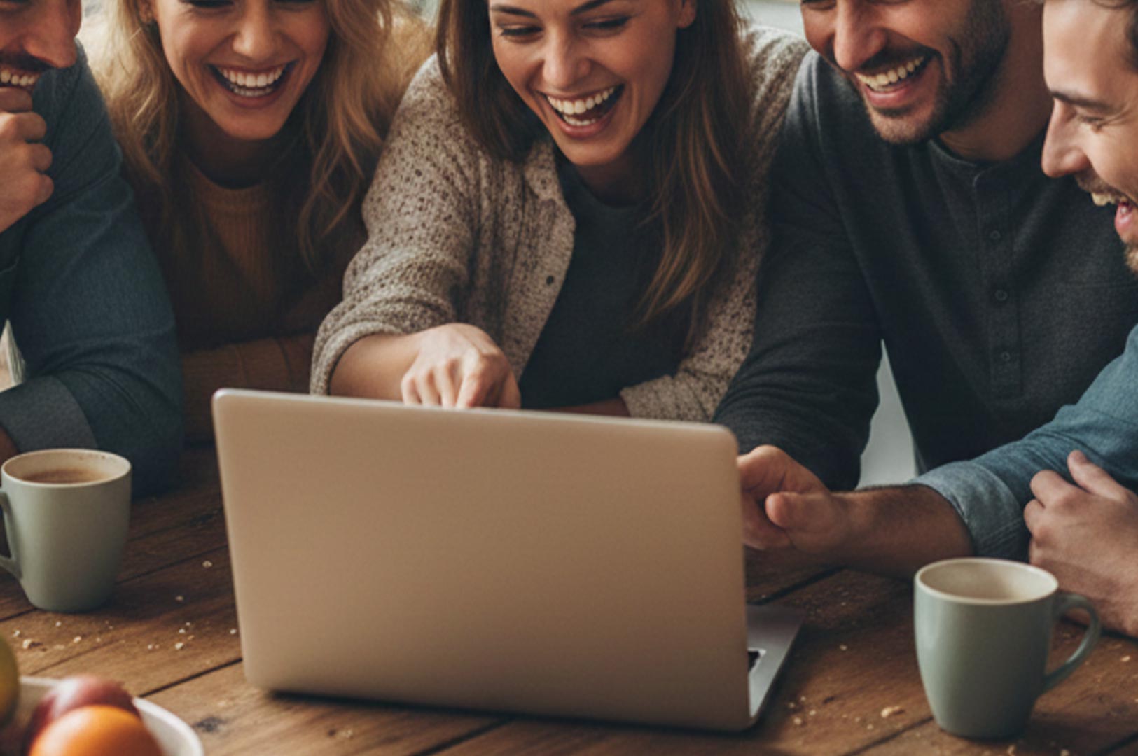 A group of happy friends gathered around a wooden table, laughing and pointing at something on a laptop screen while enjoying coffee.