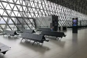 A wide-angle, eye-level shot captures a modern and spacious airport terminal gate area, characterized by a striking, massive wall of floor-to-ceiling windows supported by a complex white geometric space-frame structure of triangles and diagonals. Rows of sleek, dark gray perforated metal tandem seating units are arranged across the polished, light-gray tiled floor, all of which are currently empty, evoking a sense of stillness. To the right, a gray rectangular jet bridge entrance marked with a large number "23" stands adjacent to a small service desk and a tall, black digital information kiosk displaying a vertical screen. The bright, overcast light from outside floods the interior, emphasizing the clean lines, repetitive patterns of the architecture, and the minimalist, industrial aesthetic of the terminal.