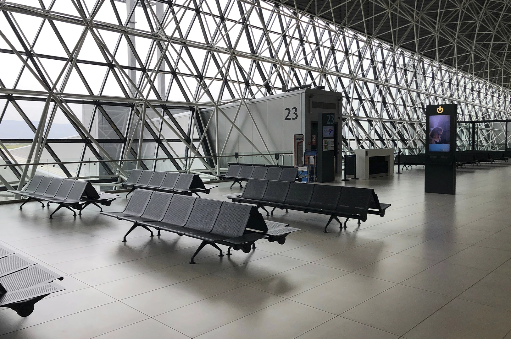 A wide-angle, eye-level shot captures a modern and spacious airport terminal gate area, characterized by a striking, massive wall of floor-to-ceiling windows supported by a complex white geometric space-frame structure of triangles and diagonals. Rows of sleek, dark gray perforated metal tandem seating units are arranged across the polished, light-gray tiled floor, all of which are currently empty, evoking a sense of stillness. To the right, a gray rectangular jet bridge entrance marked with a large number "23" stands adjacent to a small service desk and a tall, black digital information kiosk displaying a vertical screen. The bright, overcast light from outside floods the interior, emphasizing the clean lines, repetitive patterns of the architecture, and the minimalist, industrial aesthetic of the terminal.