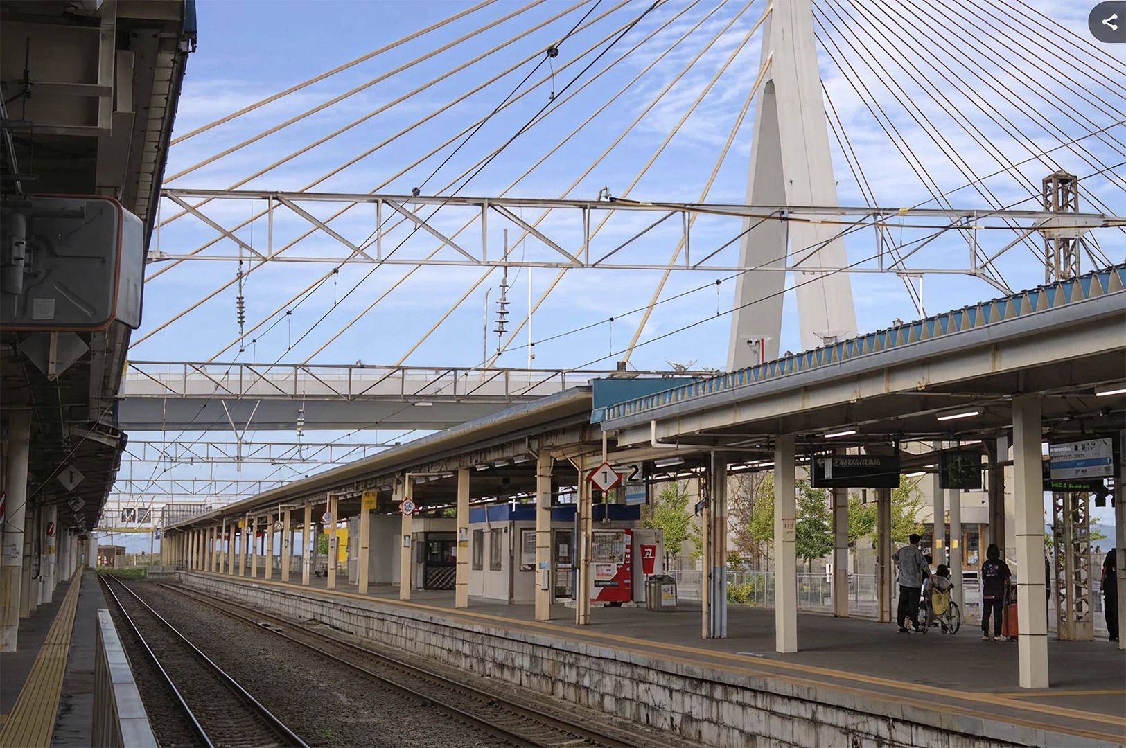 A wide-angle, eye-level shot of a quiet outdoor train platform in Japan under a bright blue sky with wispy clouds. To the right, a few commuters stand near digital displays and a red vending machine. The perspective follows the empty railway tracks receding into the distance on the left. In the background, a massive white cable-stayed bridge pylon rises high above the station’s industrial steel framework and overhead power lines.