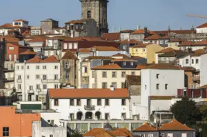 Crowded cityscape showcases a dense cluster of traditional buildings with terracotta tiled roofs and walls in varying shades of white, cream, yellow, and deep ochre. The architecture is a mix of multi-story townhouses with small rectangular windows and narrow balconies, some showing signs of age and weathering. In the background, the weathered stone bell tower of a historic church rises above the rooftops against a clear, pale blue sky. The scene is bathed in bright, direct sunlight, creating sharp shadows that emphasize the layered, hilly arrangement of the structures, while a yellow construction crane is visible on the far right horizon, suggesting a blend of old-world charm and modern development.