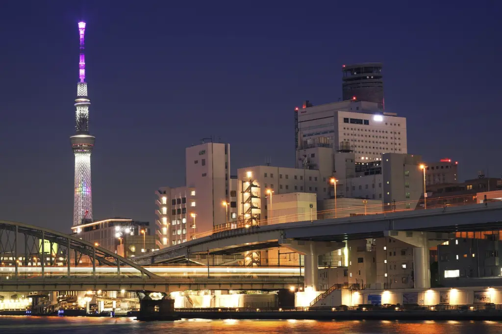 A vibrant nighttime cityscape of Tokyo featuring the Tokyo Skytree illuminated in purple and white lights. In the foreground, a multi-level concrete bridge and highway span across a river, with the long-exposure streaks of vehicle lights creating glowing horizontal lines. Several mid-rise office buildings with lit windows stand between the river and the tower under a deep twilight sky.