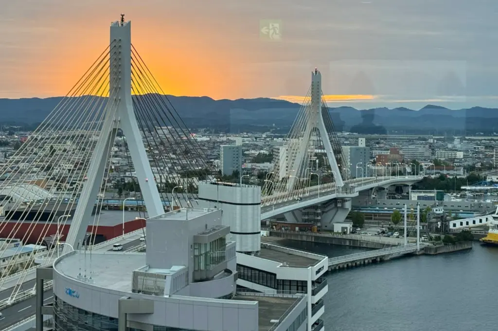A high-angle, scenic view of the Aomori Bay Bridge at sunset. The iconic white, cable-stayed bridge spans across the water, with the sun setting behind distant mountains in a warm, orange-hued sky. A modern building is in the foreground, and the harbor area stretches out beneath the bridge.