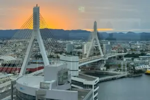 A high-angle, scenic view of the Aomori Bay Bridge at sunset. The iconic white, cable-stayed bridge spans across the water, with the sun setting behind distant mountains in a warm, orange-hued sky. A modern building is in the foreground, and the harbor area stretches out beneath the bridge.