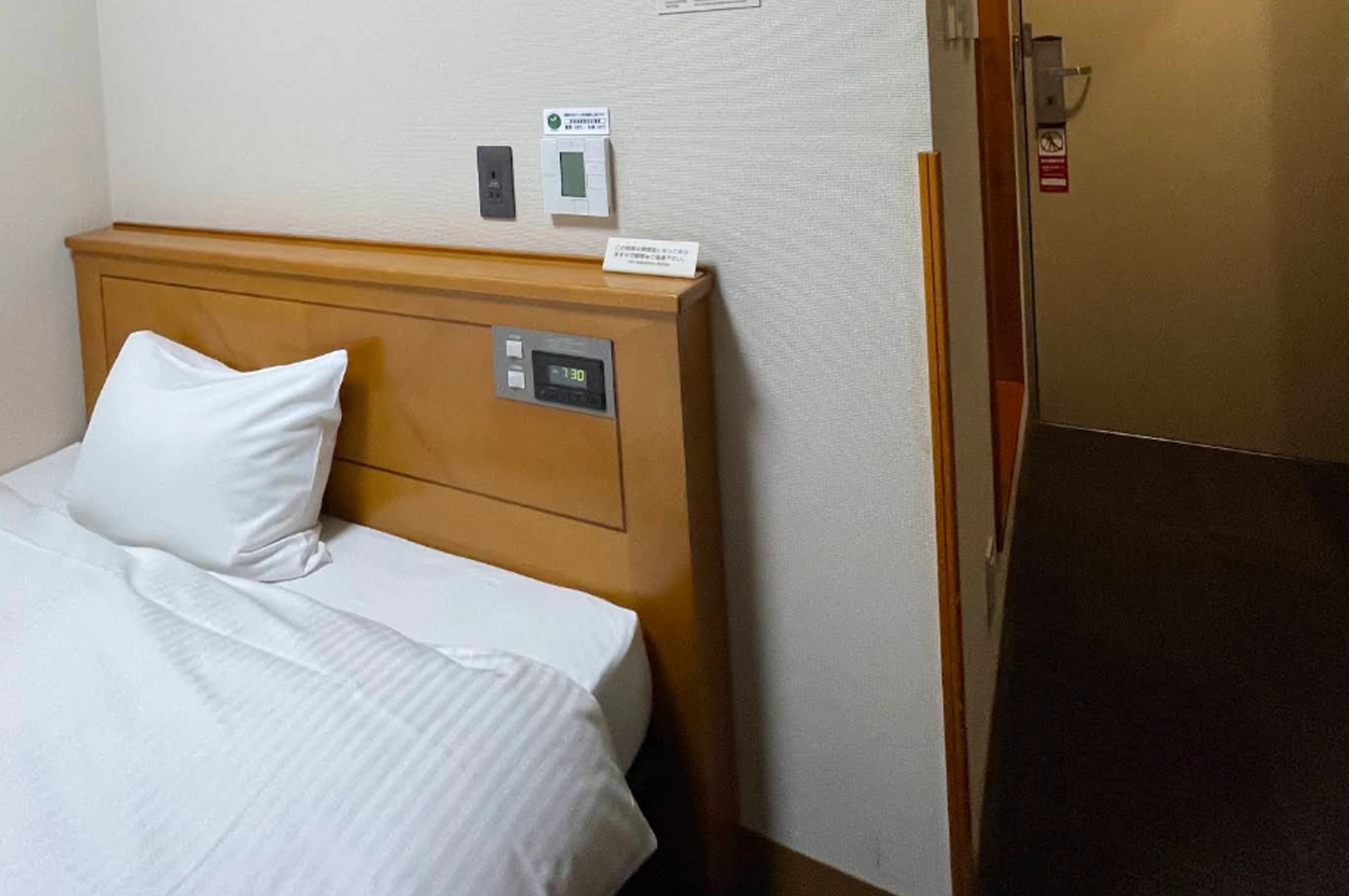 A clean, minimalist Japanese hotel room featuring a single bed with white linens and a light wood headboard. Integrated into the headboard is a digital clock showing "7:30" and light switches. A white climate control panel and a black power outlet are mounted on the wall above.