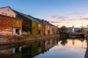 A serene sunset view of the Otaru Canal in Hokkaido, Japan, featuring ivy-covered stone warehouses reflected in the water and a glowing lighthouse in the distance.