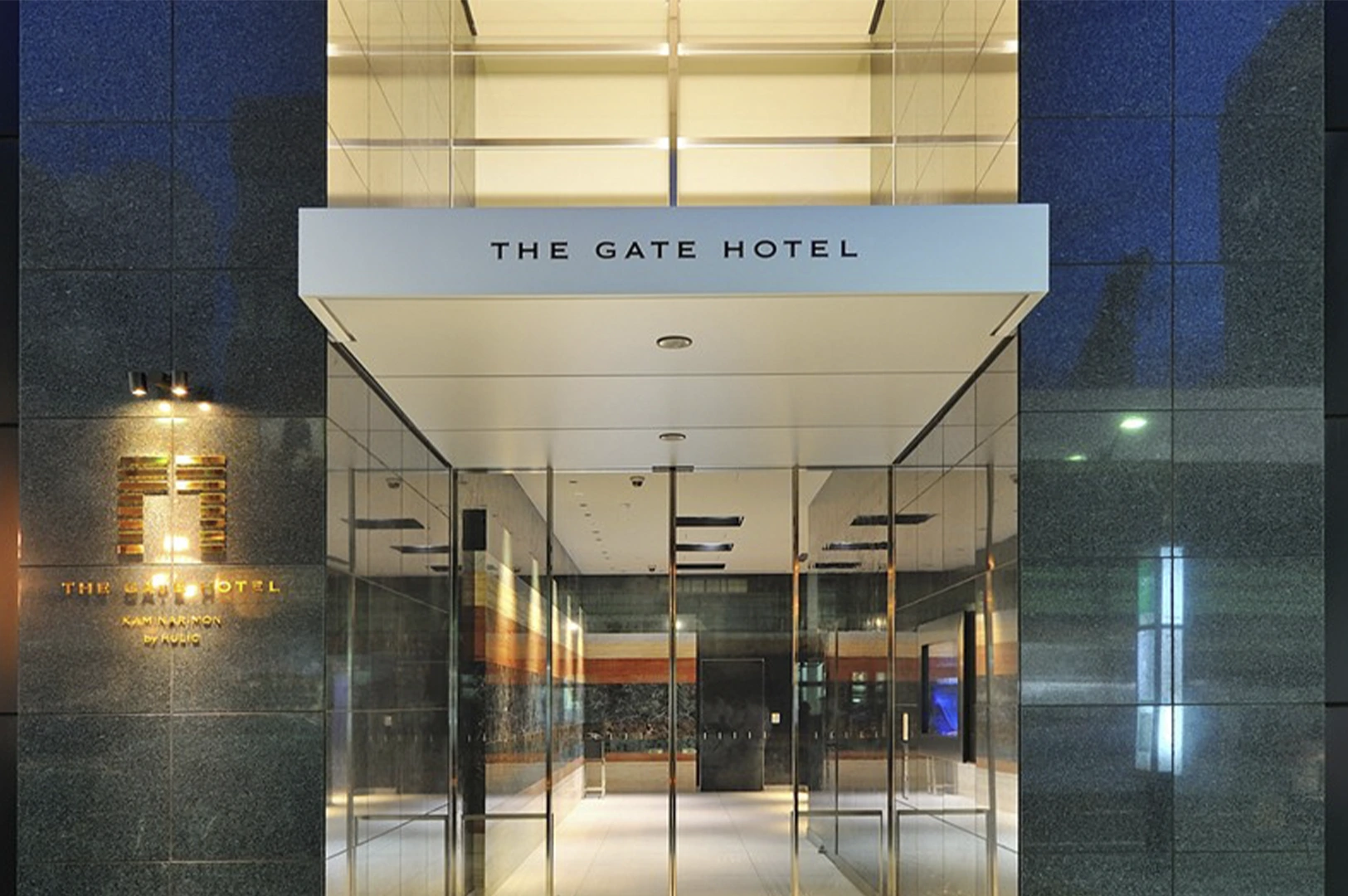 A sophisticated, symmetrical view of the entrance to The Gate Hotel Kaminarimon by Hulic. The entrance is flanked by dark, polished emerald-green marble walls. A white rectangular canopy with the hotel's name hangs over the glass doors. To the left, a gold-embossed logo is illuminated on the marble wall, and the interior hallway features warm, horizontal-striped stone walls.