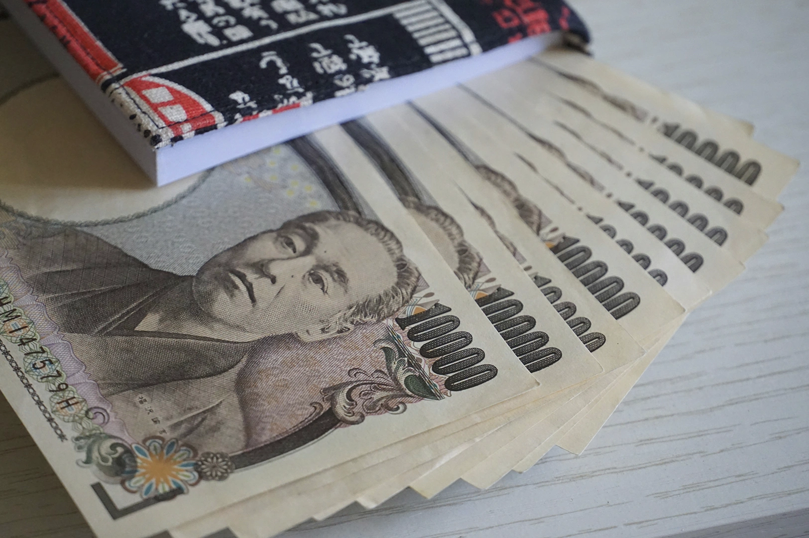 A close-up, slightly angled shot of a stack of Japanese 10,000 yen banknotes fanned out on a table next to a small, patterned fabric notebook or wallet. The face of Yukichi Fukuzawa is clearly visible on the currency.