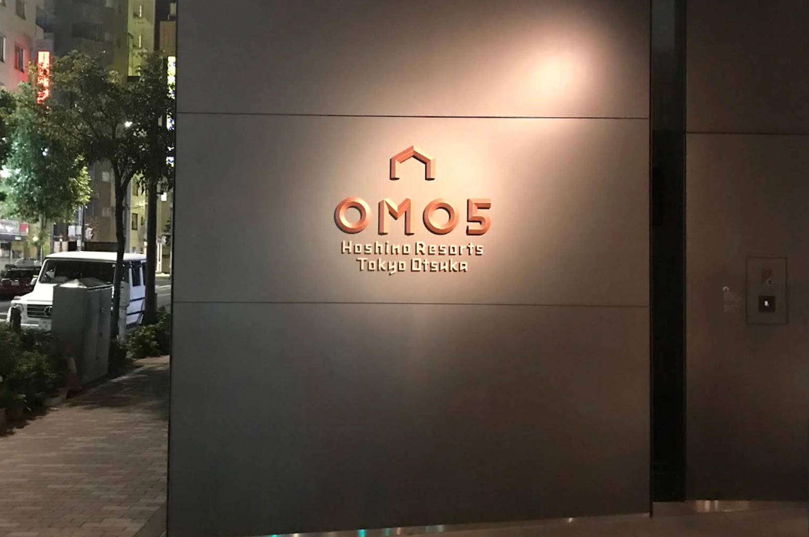 A close-up shot of an exterior wall at night featuring the illuminated logo for OMO5 Tokyo Otsuka by Hoshino Resorts. The wall consists of large, smooth grey panels with a matte finish. The logo and text are rendered in a warm, copper-toned metallic material that glows under a soft overhead spotlight. Above the bold "OMO5" text is a minimalist icon of a house or roof. A portion of a brick sidewalk, a green tree, and a white van parked on a city street are visible in the blurred background to the left.
