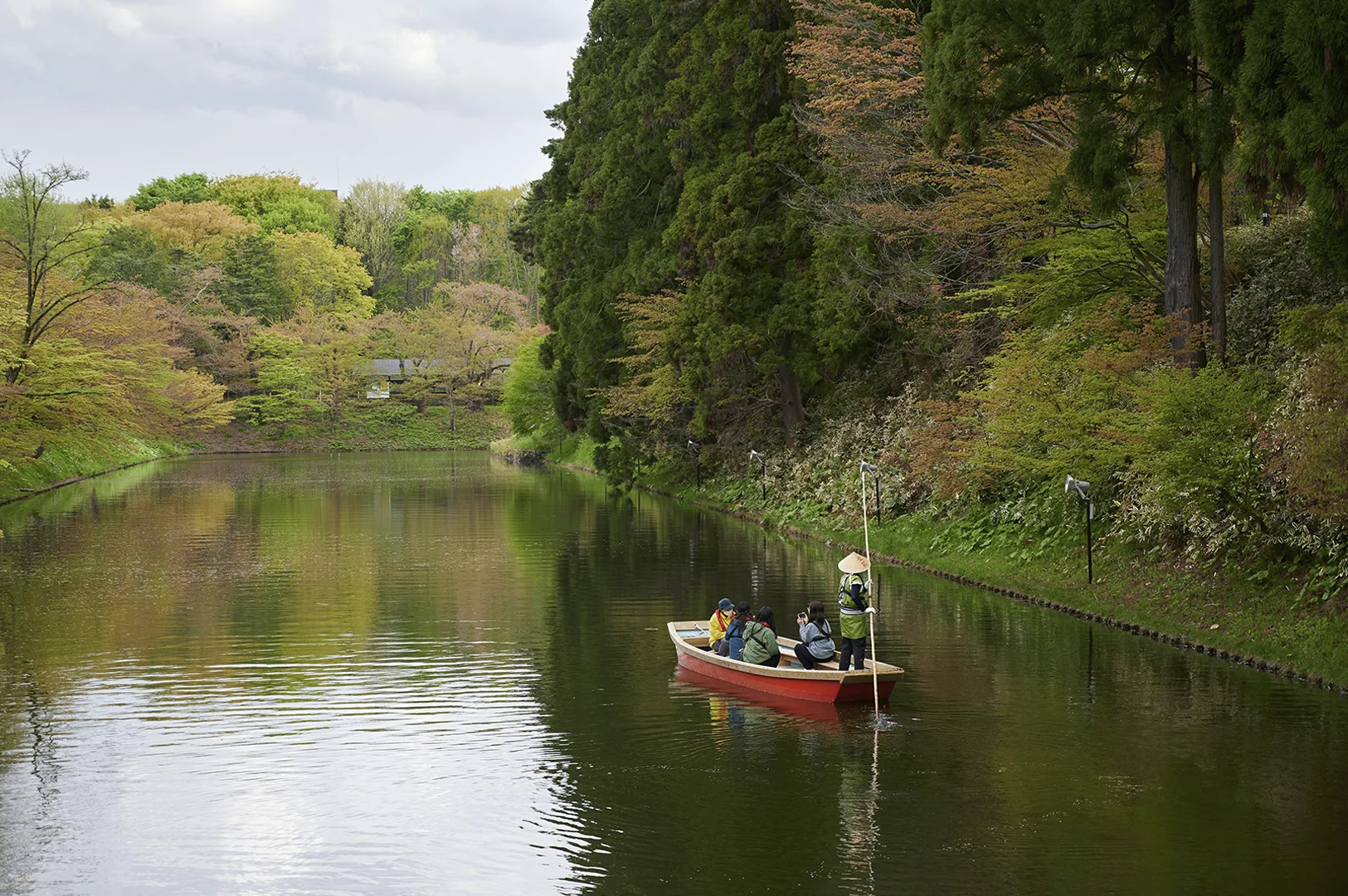 A high-angle landscape shot of a traditional flat-bottomed wooden boat being poled down a calm, dark green river or moat. The boat carries four passengers and a guide wearing a traditional conical straw hat. The water is flanked by steep, lush embankments covered in dense green trees and light-colored blossoms. The reflection of the vibrant foliage ripples across the water’s surface under an overcast sky.