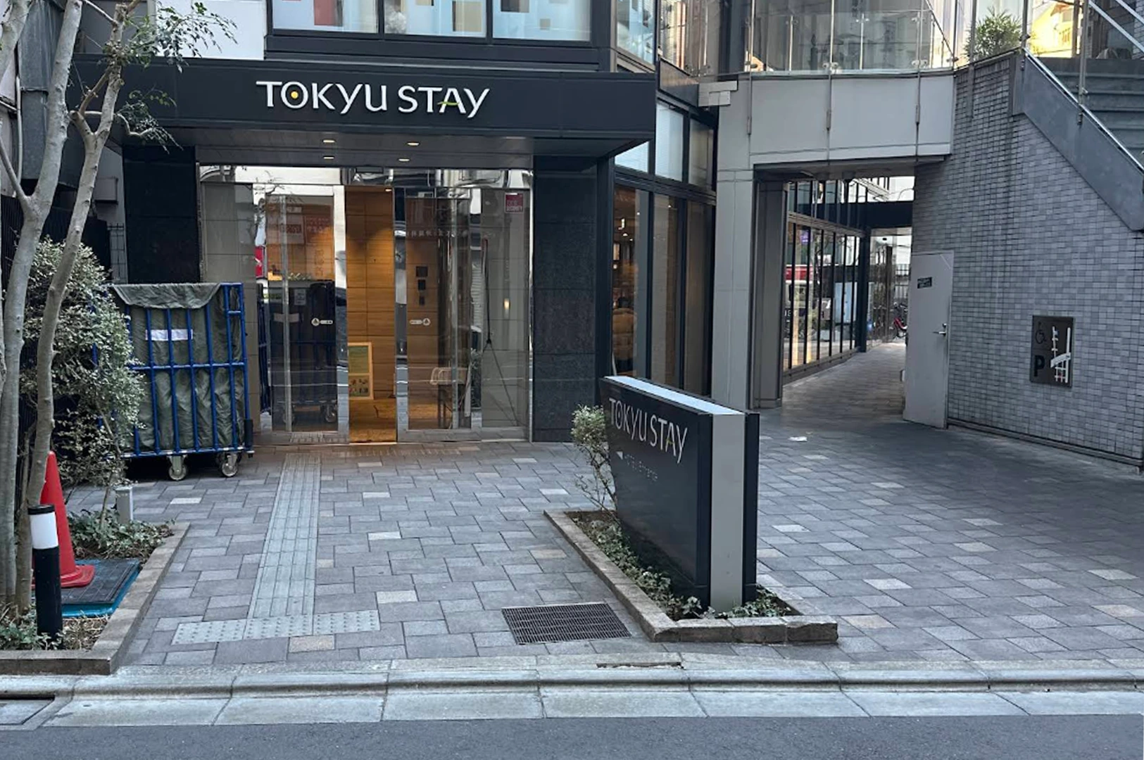 The street-level entrance of a Tokyu Stay hotel. The facade features dark grey panels with the hotel name in white and yellow lettering. A stone-paved walkway leads to glass sliding doors. To the left, a blue luggage cart and a red traffic cone are visible near a small tree, while a narrow alleyway extends to the right of the building.