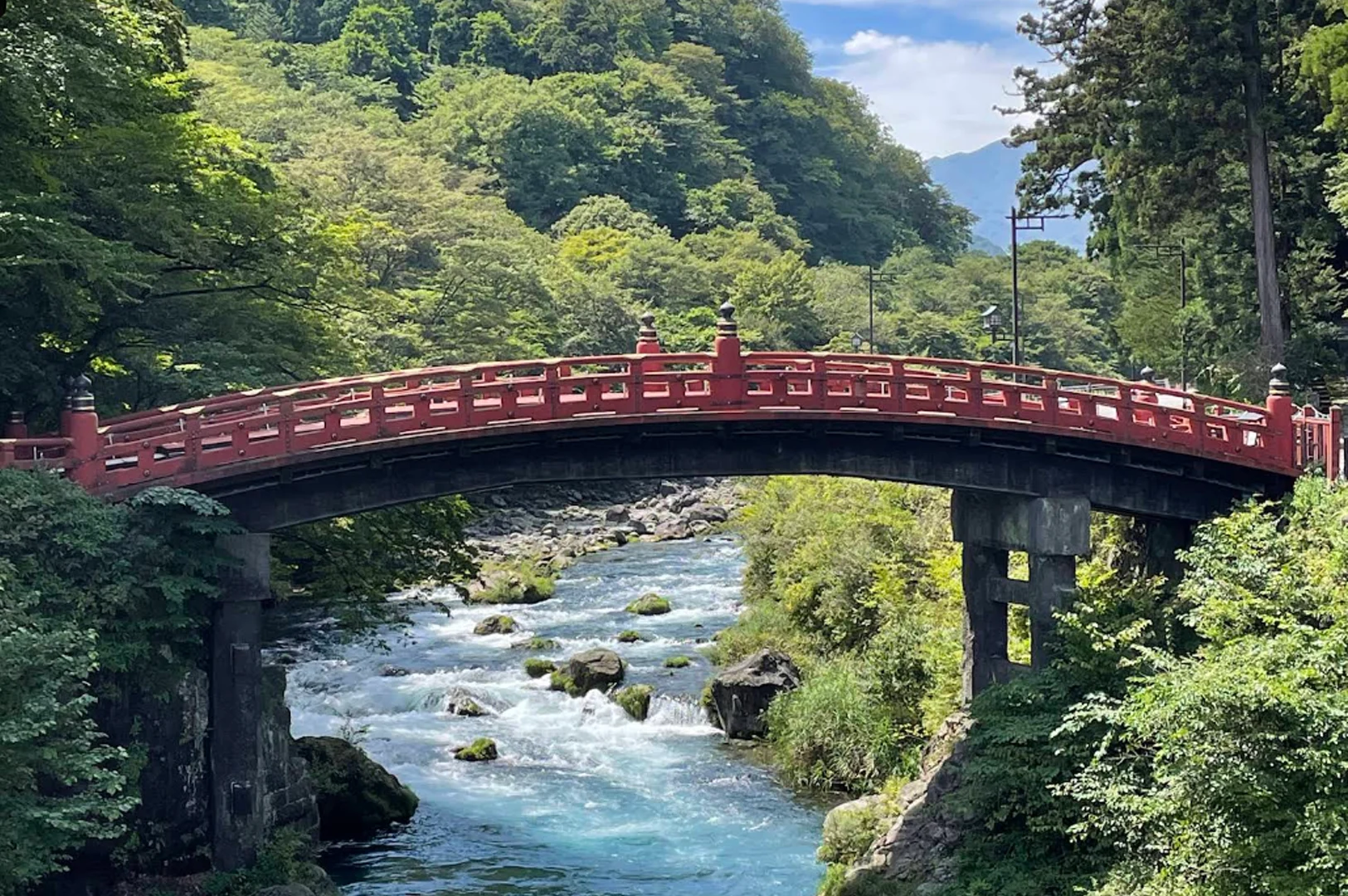 A wide-angle shot of the iconic Shinkyo Bridge, a striking vermilion-lacquered wooden bridge with black accents. It arches gracefully over a rushing river filled with turquoise water and grey boulders. The surrounding embankments are lush with thick, vibrant green summer foliage and tall evergreen trees. The scene is bright and peaceful, showcasing the contrast between the red bridge and the cool blue water.