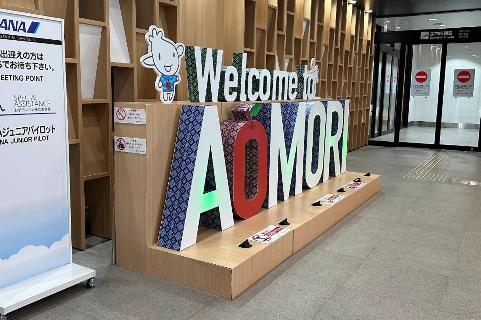 A brightly lit, welcoming display sign at an airport terminal that reads "Welcome to AOMORI" in large, colorful, 3D block letters. The letter "O" is shaped like a red apple. To the left, there is a tall informational sign for ANA (All Nippon Airways) featuring a cartoon mascot.