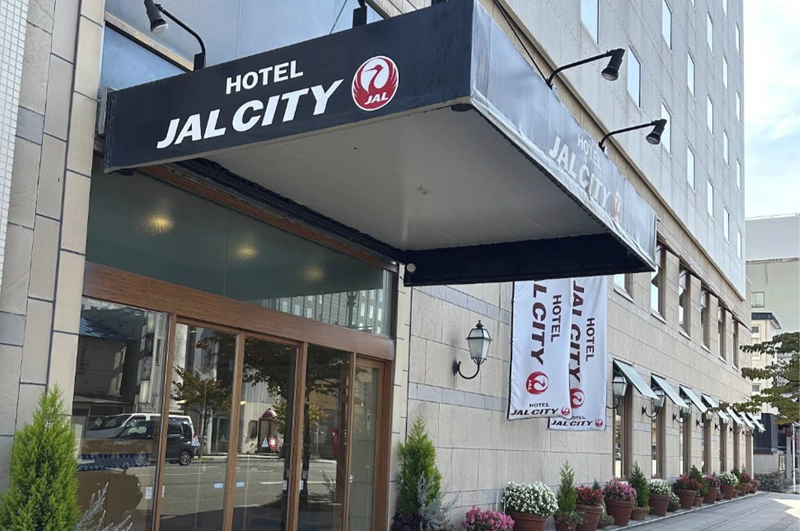 The exterior entrance of Hotel JAL City. A large black rectangular awning with white lettering and the red JAL logo extends over the glass doors. Matching white banners hang from the stone facade, and several potted pink and white flowers line the sidewalk.