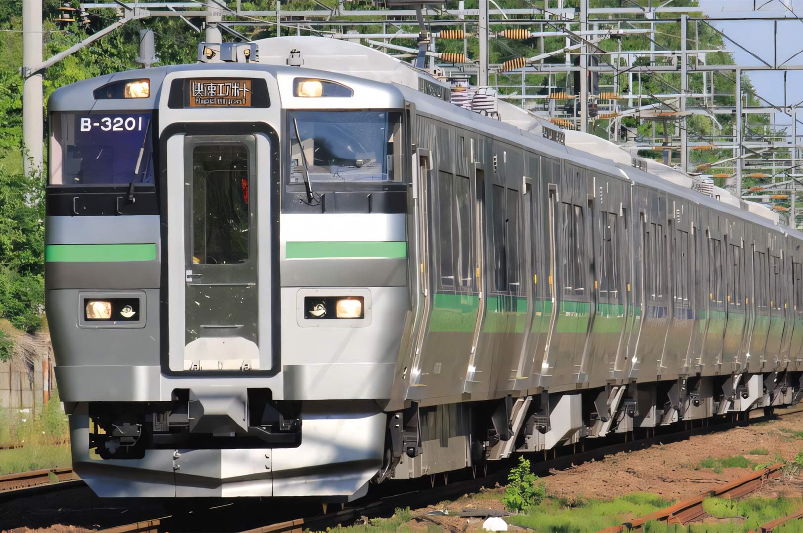A silver JR Hokkaido 733 series commuter train (B-3201) with green stripes traveling along a railway track in Japan during the day.