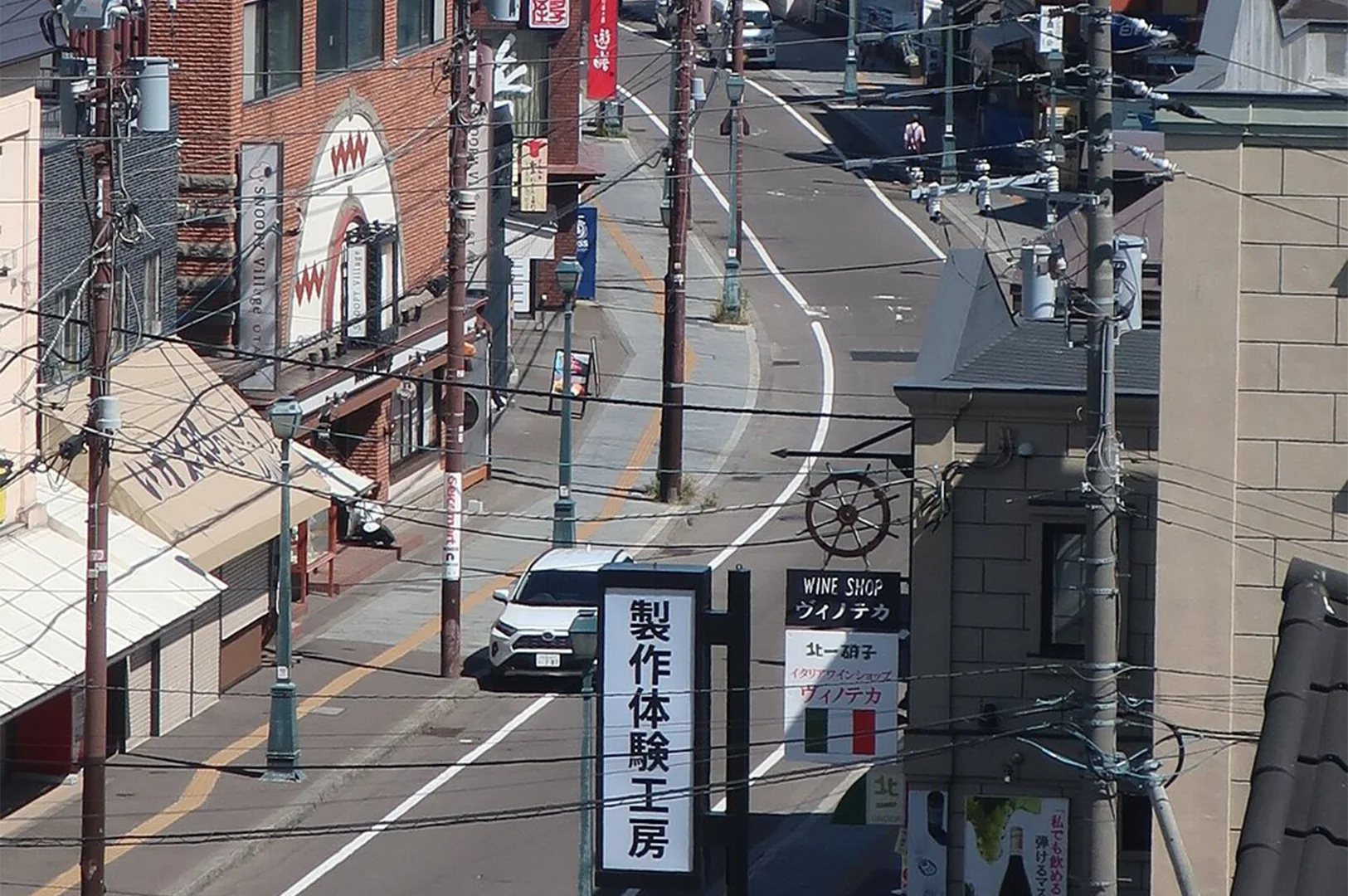 A high-angle view of a winding city street in Otaru, Japan, showing local shops, a white car driving past Japanese signage, and a network of overhead power lines.