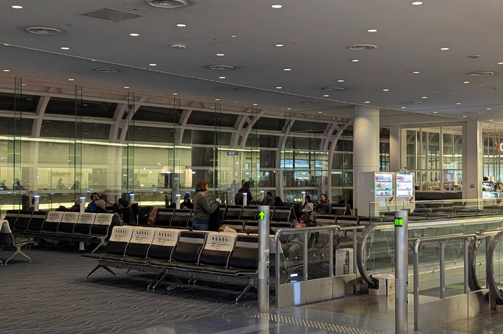 An interior shot of a spacious, modern airport departure lounge at night. Rows of black upholstered seating with white headrest covers are mostly empty, with a few travelers scattered throughout. Large floor-to-ceiling glass windows reveal the tarmac and airport structures outside. To the right, a moving walkway with glowing green directional arrows sits next to a glass partition and a illuminated vending machine.