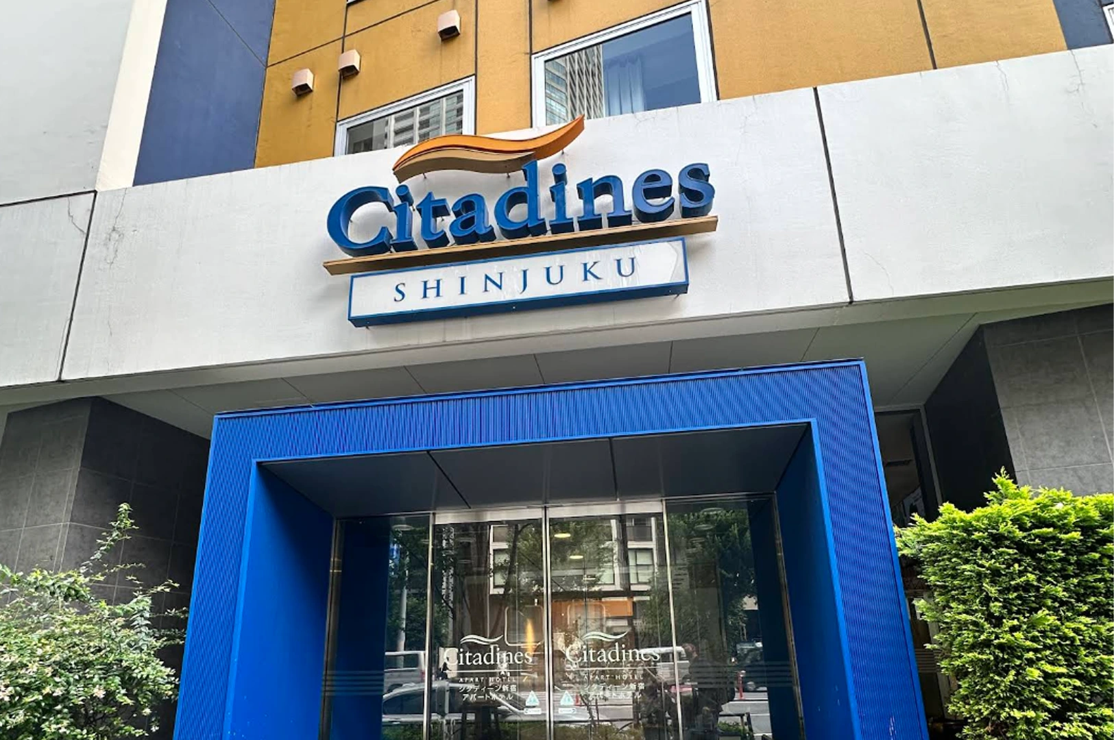 A close-up shot of the entrance to the Citadines Shinjuku apartment hotel. The building has a white and ochre-yellow exterior. A large blue sign with "Citadines" in stylized script sits above a smaller "SHINJUKU" sign. The main doorway is framed by a bright blue, rectangular architectural portico with glass doors reflecting the street.