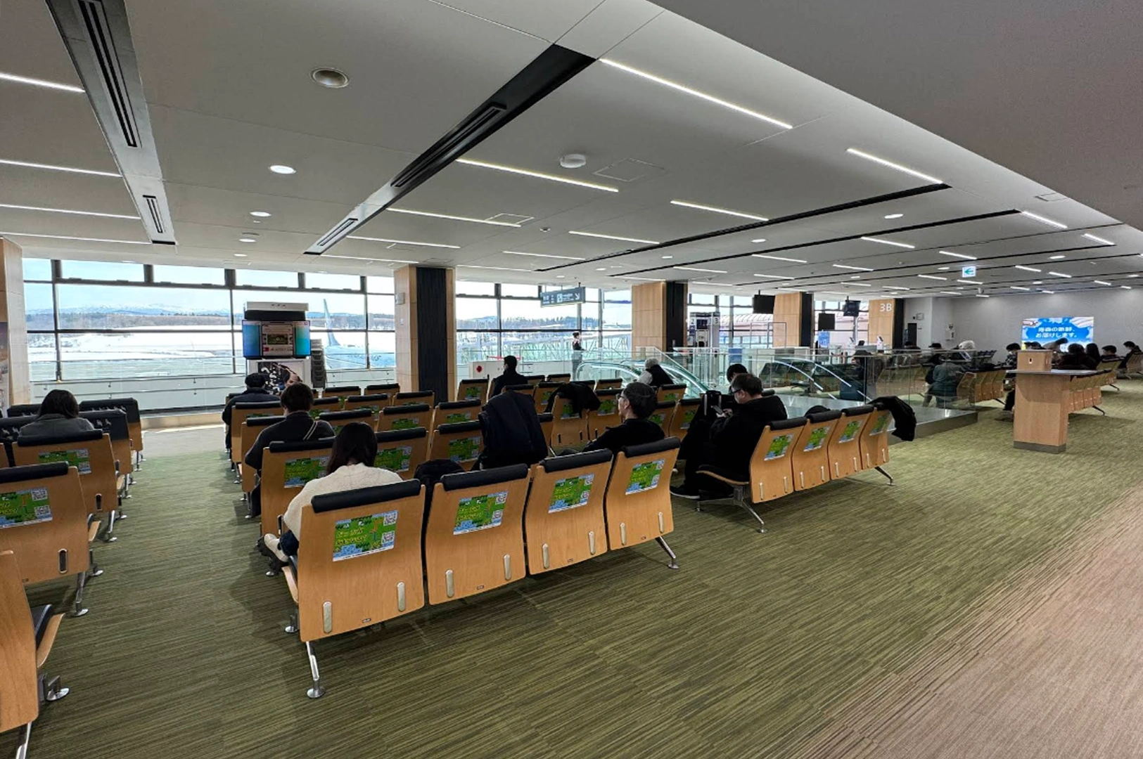 An interior view of a quiet, modern airport waiting lounge. Rows of wooden-backed chairs with black cushions and green informational stickers face large floor-to-ceiling windows. Through the glass, a snowy tarmac is visible under a bright, overcast sky, with the tail of a blue airplane parked at a gate. The room features clean lines, recessed LED lighting, and polished green-toned carpeting, creating a calm, minimalist atmosphere.