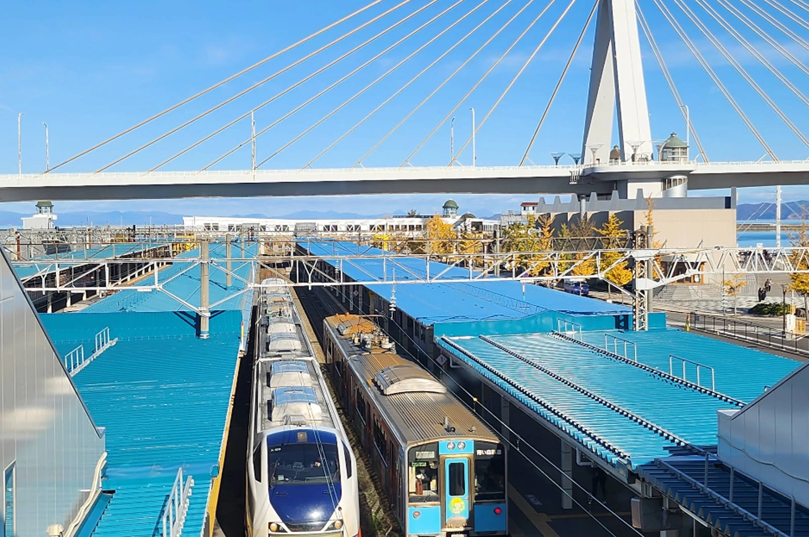 A high-angle view of Aomori Station in Japan under a clear blue sky. Several trains are parked at the platforms beneath vibrant blue roofing. In the background, the large white cables and pylon of the Aomori Bay Bridge span across the frame.