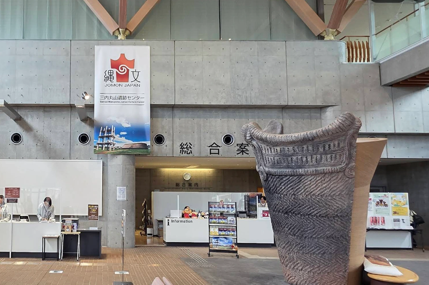 An indoor shot of the entrance to the Sannai-Maruyama Jomon Culture Center. In the foreground, a large, textured dark gray replica of a prehistoric Jomon pottery vessel stands prominently. Behind it, a clean, industrial-style lobby features a concrete wall with a large banner displaying a reconstructed wooden structure and the "Jomon Japan" logo. Staff members in light-colored uniforms work behind white reception desks.