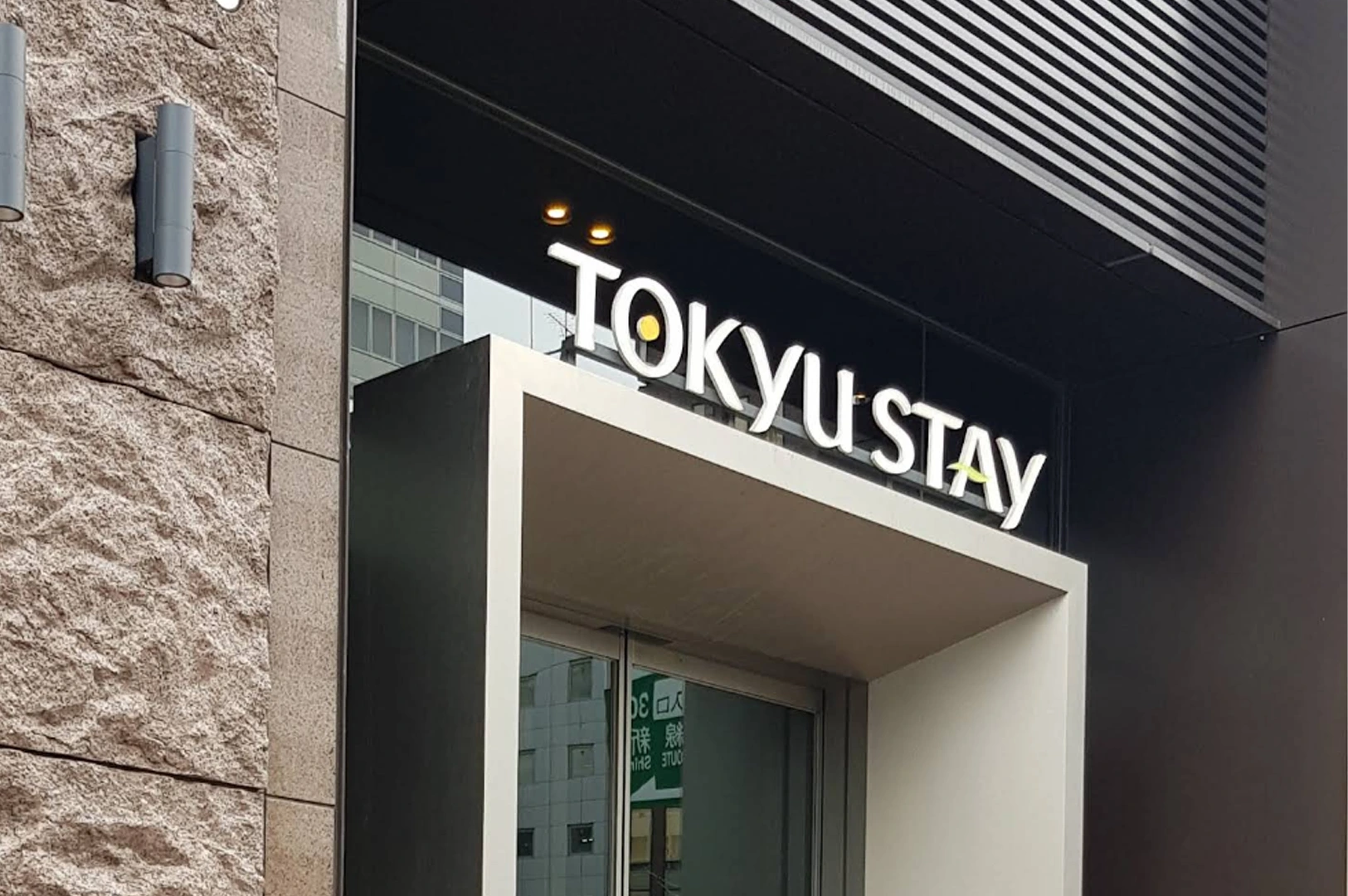 A low-angle, close-up shot of a Tokyu Stay hotel sign. The white illuminated letters are mounted on a dark horizontal beam above a modern, cream-colored rectangular entryway. To the left, the building's exterior is finished with a rough-textured, light-brown stone tile and two minimalist black cylindrical wall lamps.