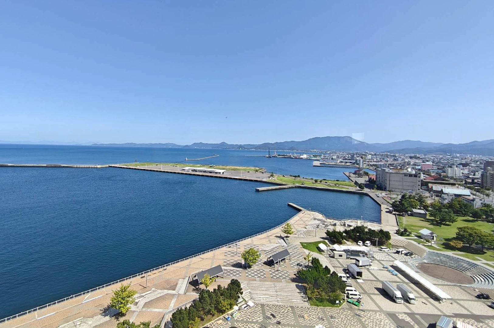 A wide, elevated daytime view of the Aomori coastline. A large, deep blue bay stretches out toward the horizon, meeting a clear blue sky. In the foreground, a paved plaza and harbor area lead to the water's edge, with distant city buildings and mountains visible in the background.
