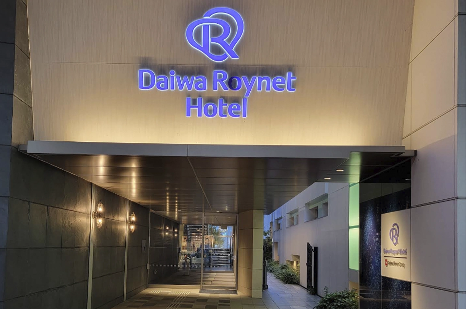 The nighttime entrance of a Daiwa Roynet Hotel. A large, glowing blue logo and "Daiwa Roynet Hotel" text are illuminated above a modern, recessed entryway with dark stone walls and warm overhead lighting.