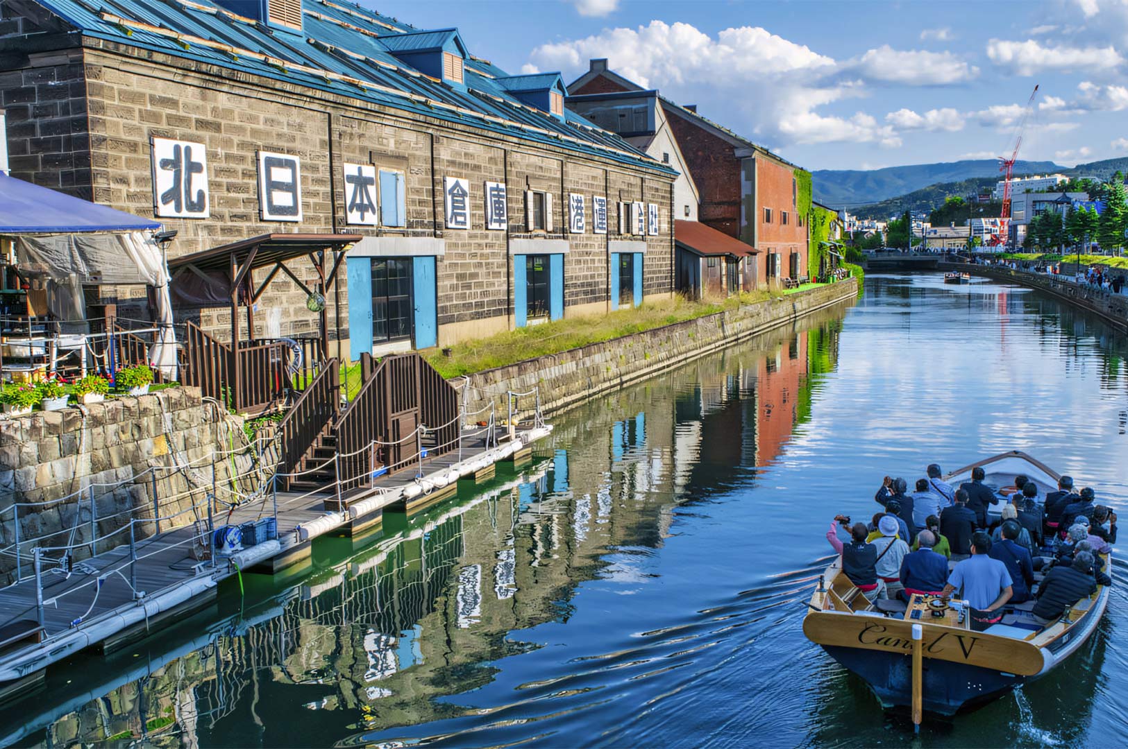 A tourist sightseeing boat cruising through the blue waters of Otaru Canal, passed historic stone buildings with Japanese signage under a bright sky.