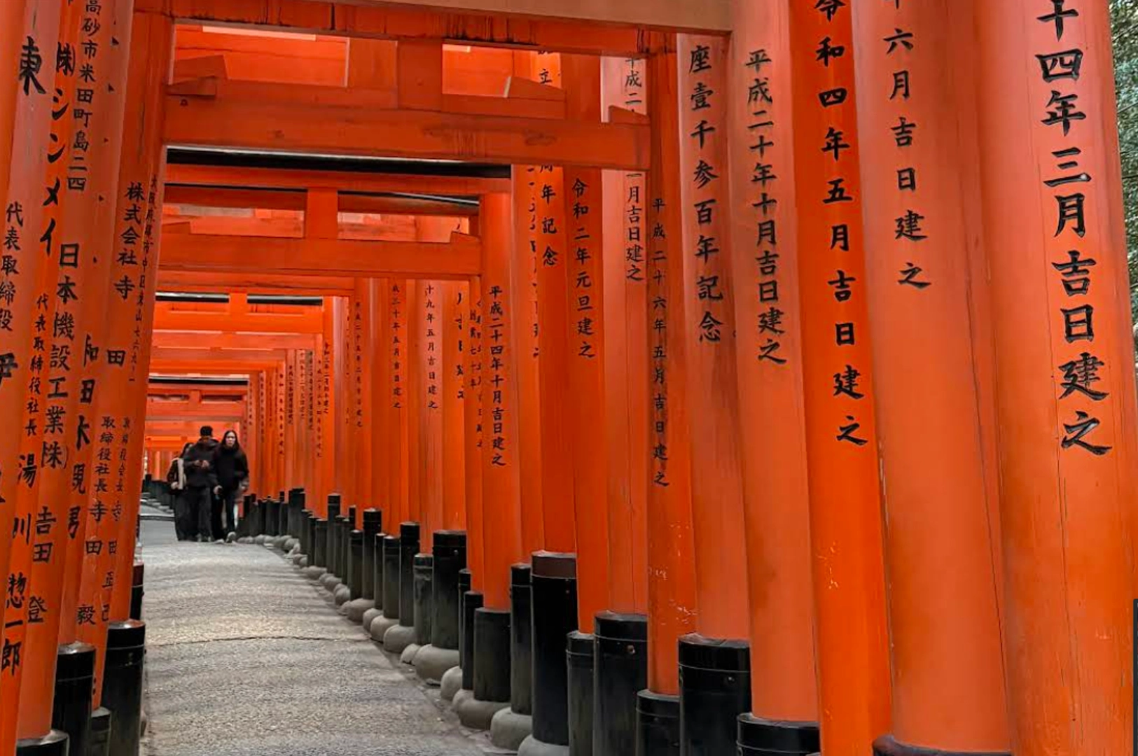 A ground-level perspective looking through a dense, receding tunnel of bright vermillion Torii gates at Fushimi Inari-taisha. The thick wooden pillars are inscribed with black Japanese characters. Two figures are visible as small silhouettes in the distance at the end of the stone path. The lighting creates a rhythmic play of shadows and saturated orange-red tones throughout the walkway.