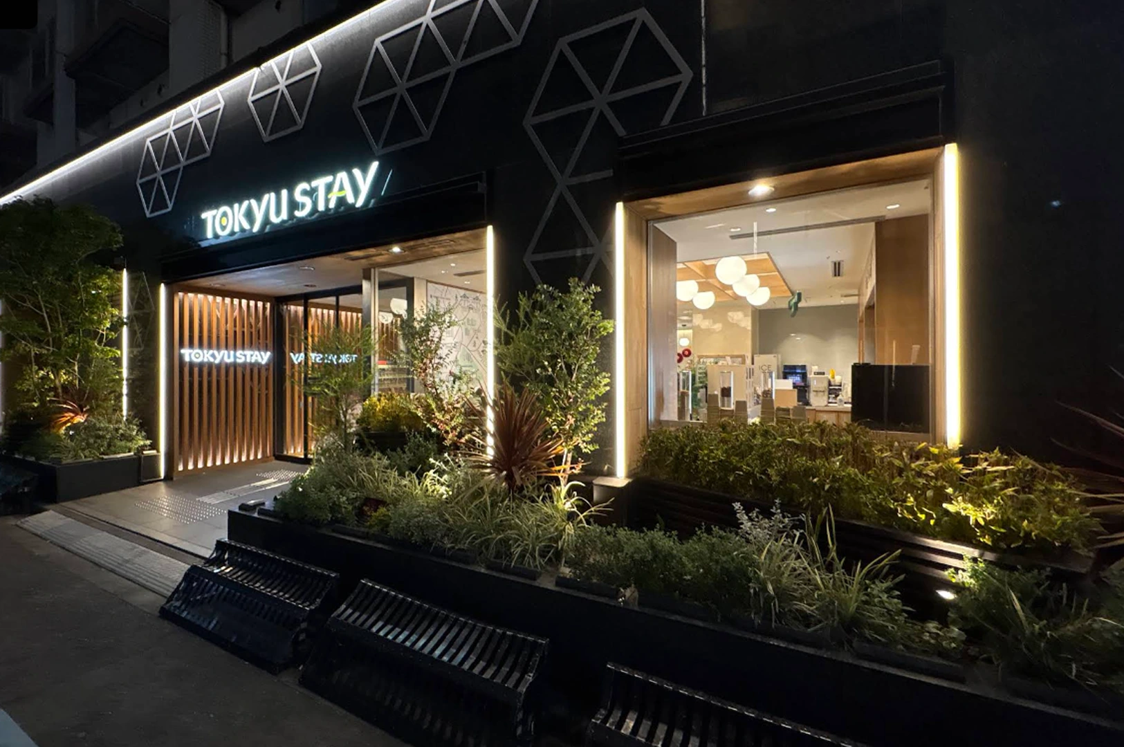 A modern hotel storefront for Tokyu Stay at night. The black facade is decorated with glowing white geometric "honeycomb" patterns. Bright vertical LED light strips frame the large glass windows and the wooden-slatted entrance. In the foreground, there are dark planters with lush greenery and several black metal benches.