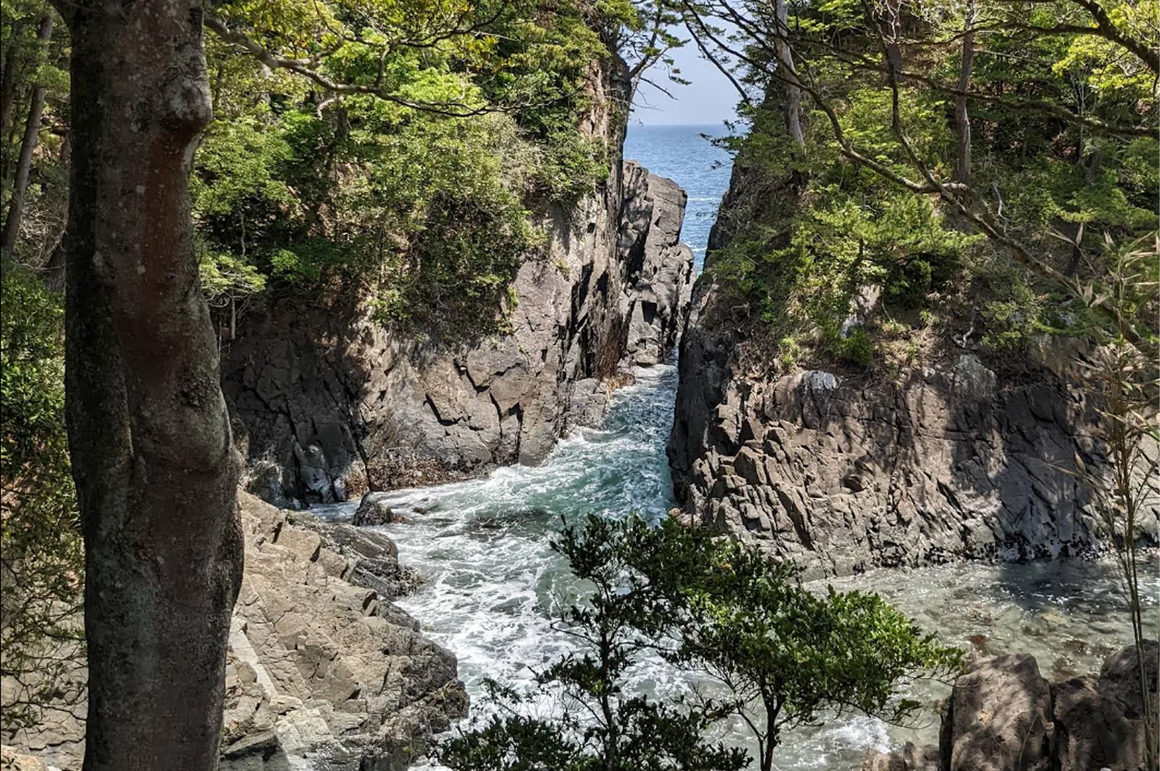 A dramatic vertical view looking through a narrow, jagged rock gorge toward the open sea. The steep, dark grey stone cliffs are topped with green trees and hardy shrubs. White-capped waves crash against the base of the rocks, sending sea foam into the narrow channel. The deep blue of the ocean meets a pale, clear sky at the horizon, framed perfectly by the natural stone walls.