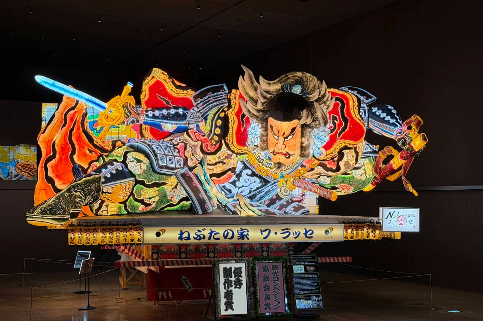 A large, intricate, and colorful Nebuta float on display inside a dimly lit museum. The float depicts a traditional Japanese warrior figure with a fierce expression, dressed in elaborate armor and holding a glowing blue sword. A sign below the float in Japanese reads "Nebuta-no-ie Warasse."