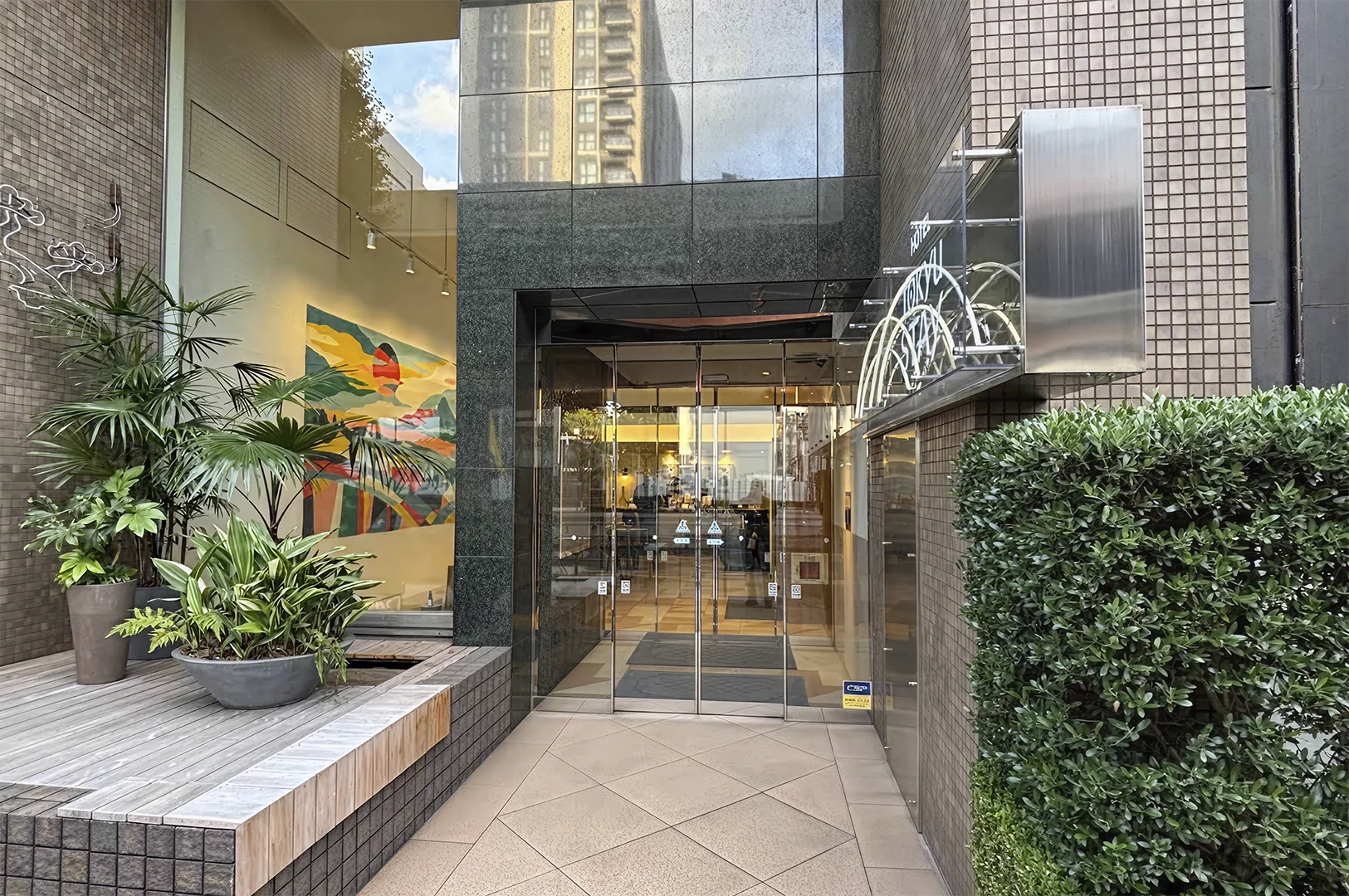 The entrance of a Tokyu Stay hotel featuring a mix of textures. The building is clad in small brown tiles and dark polished granite. Glass sliding doors lead into a brightly lit lobby. To the left of the door, a large, colorful mural depicting a stylized landscape is visible behind a glass partition, fronted by potted tropical plants on a wooden deck.