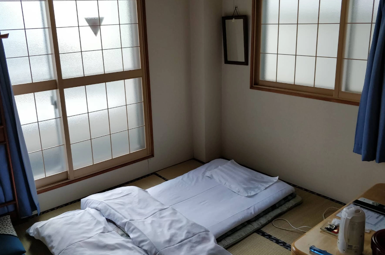 A simple and serene interior of a traditional Japanese room (washitsu). A white futon bed is neatly laid out on a beige tatami mat floor. The room features shoji screen windows with wooden frames and blue privacy curtains. In the corner, a low wooden table holds a white electric kettle and tea supplies. The lighting is soft and natural, emphasizing the minimalist and cozy nature of the accommodation.