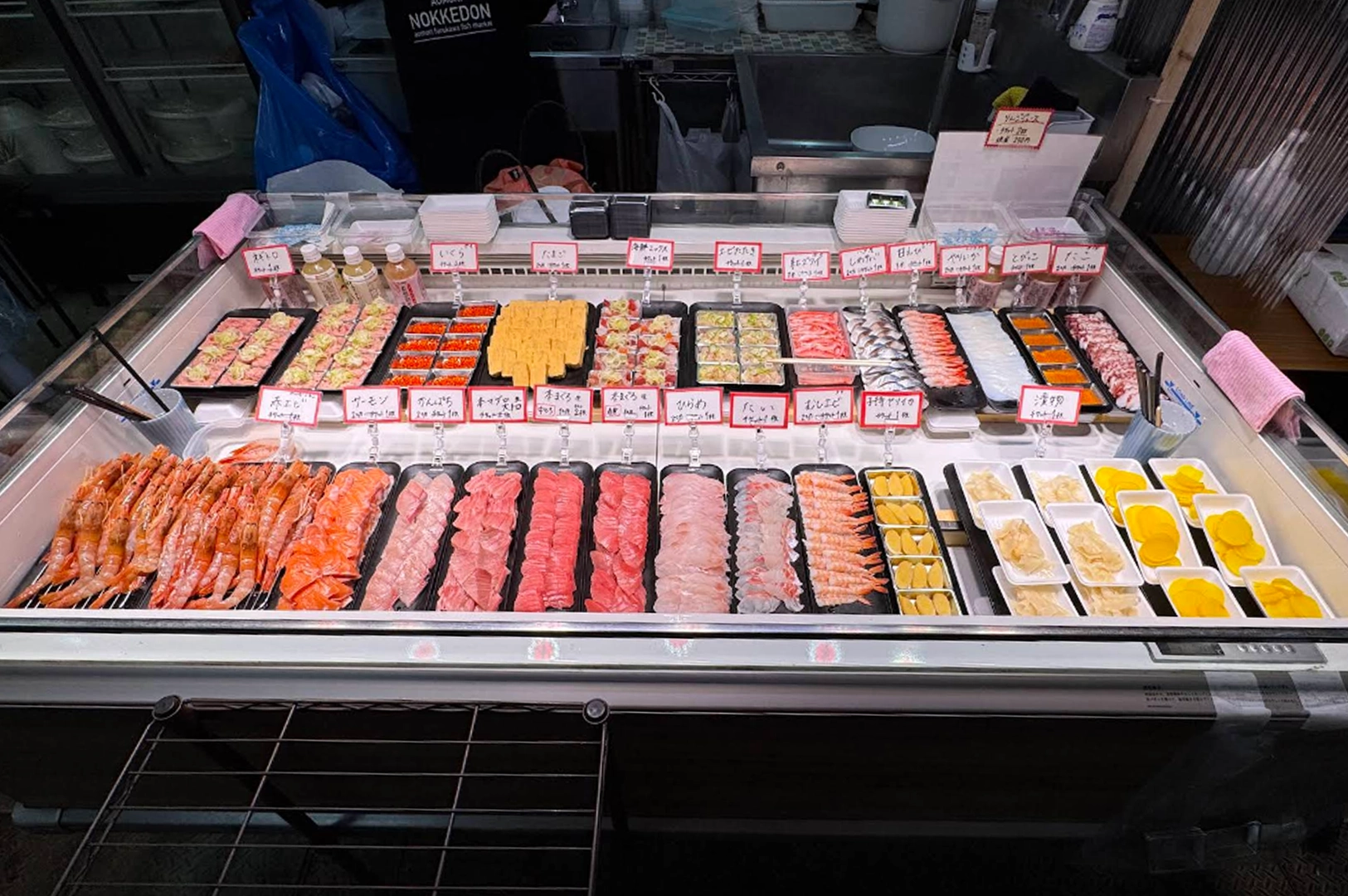 A vibrant, well-stocked refrigerated display case at a market, likely for Nokkedon (customizable seafood bowls). Various types of fresh sashimi, including slices of tuna, salmon, white fish, and rows of shrimp, are neatly arranged on black trays. Small, handwritten signs in Japanese label each seafood option.