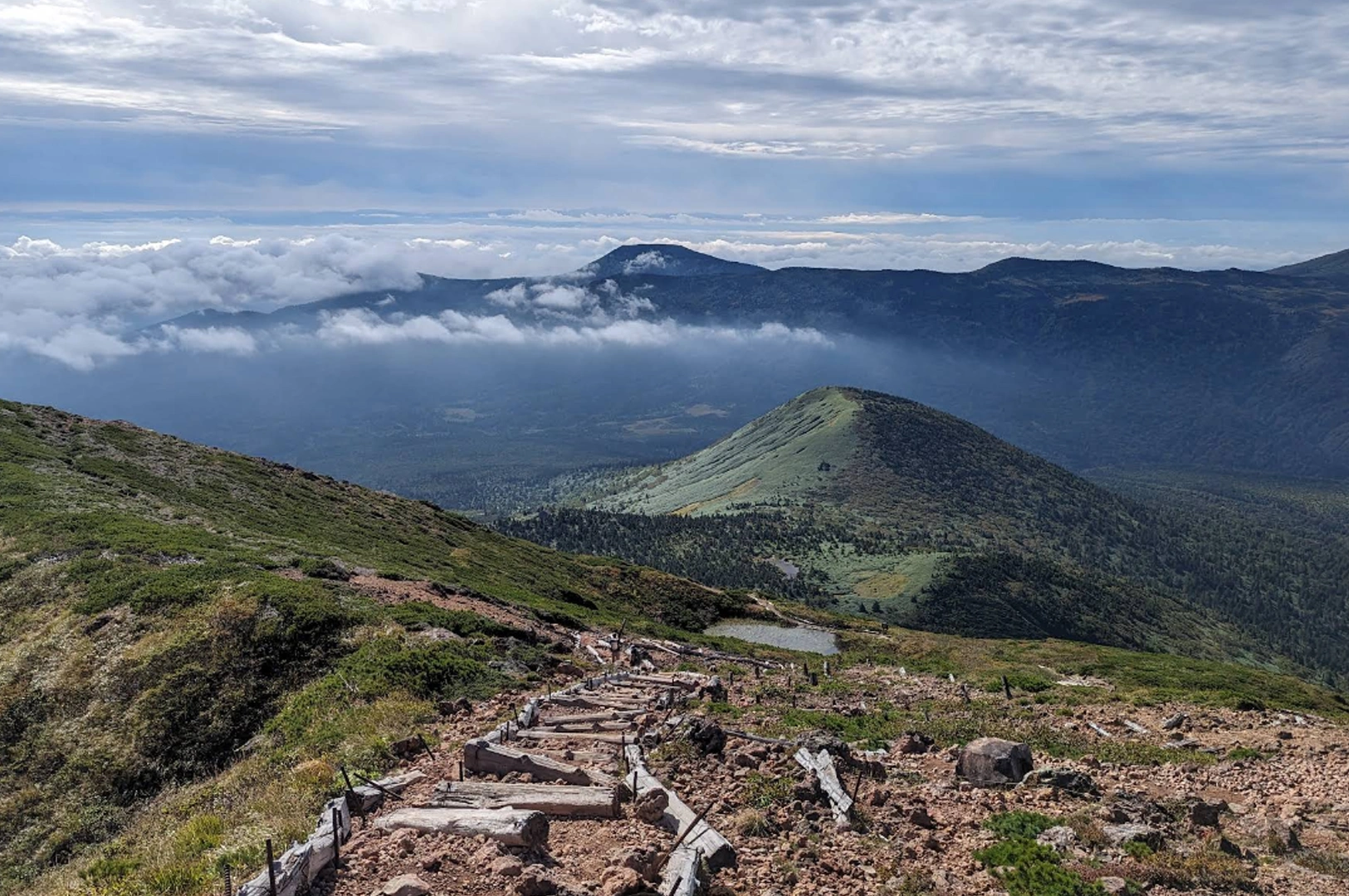 A breathtaking panoramic view from a mountain hiking trail. A rugged path of wooden steps and dirt leads the eye down toward rolling green hills and a distant, cloud-covered valley. Small alpine ponds and patches of evergreens dot the landscape.