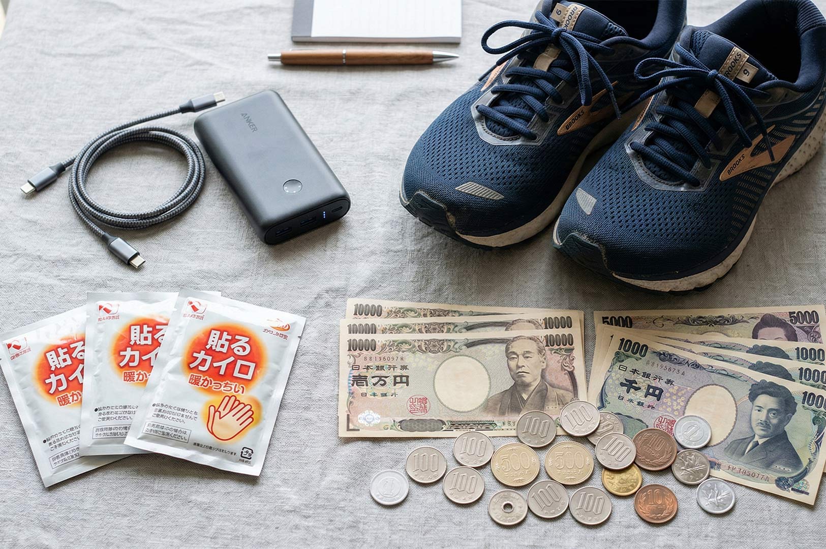 Flat lay of Japan travel essentials including Japanese Yen (10,000, 5,000, 1,000 bills and coins), Brooks running shoes, a power bank, and heat packs (kairo).