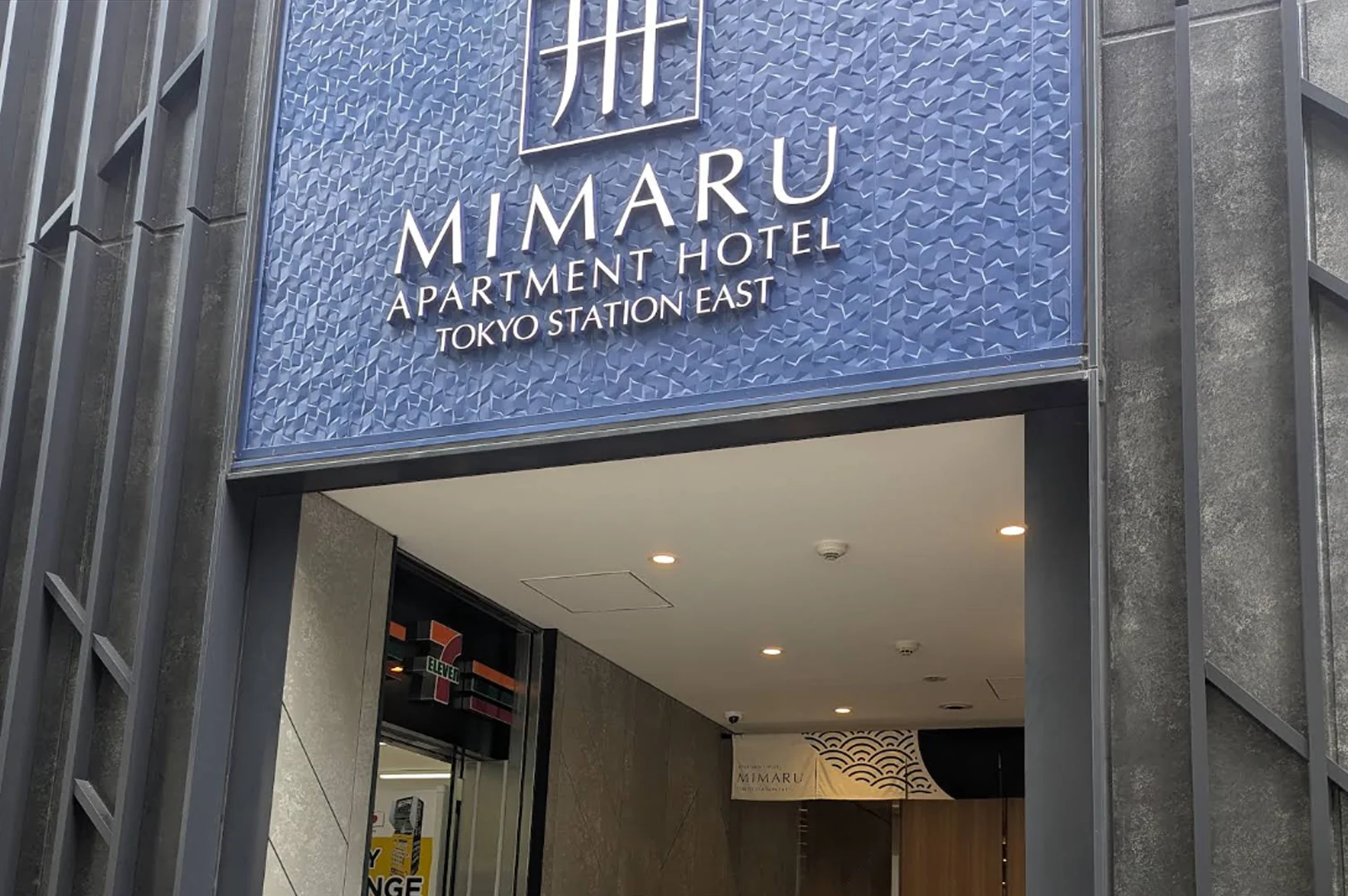 A close-up of the blue textured signage for MIMARU Apartment Hotel Tokyo Station East. The sign features a silver logo and clean white typography against a geometric, 3D-patterned blue background. Below the sign, the entryway reveals a glimpse of a 7-Eleven store and a traditional Japanese "Noren" curtain hanging in the hallway.