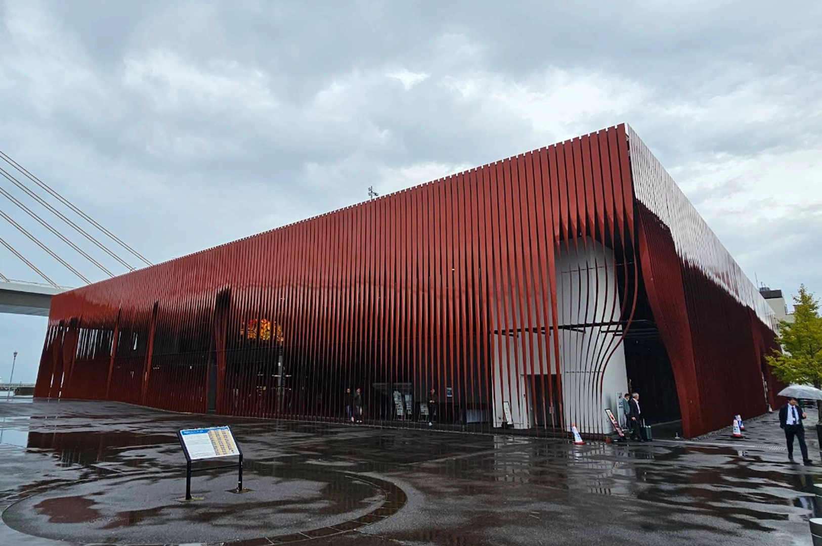 The Nebuta Museum Wa Rasse in Aomori, a striking modern building wrapped in tall, thin red metal slats that curve at the bottom to create entryways. The ground is wet from rain, reflecting the building and the overcast gray sky.