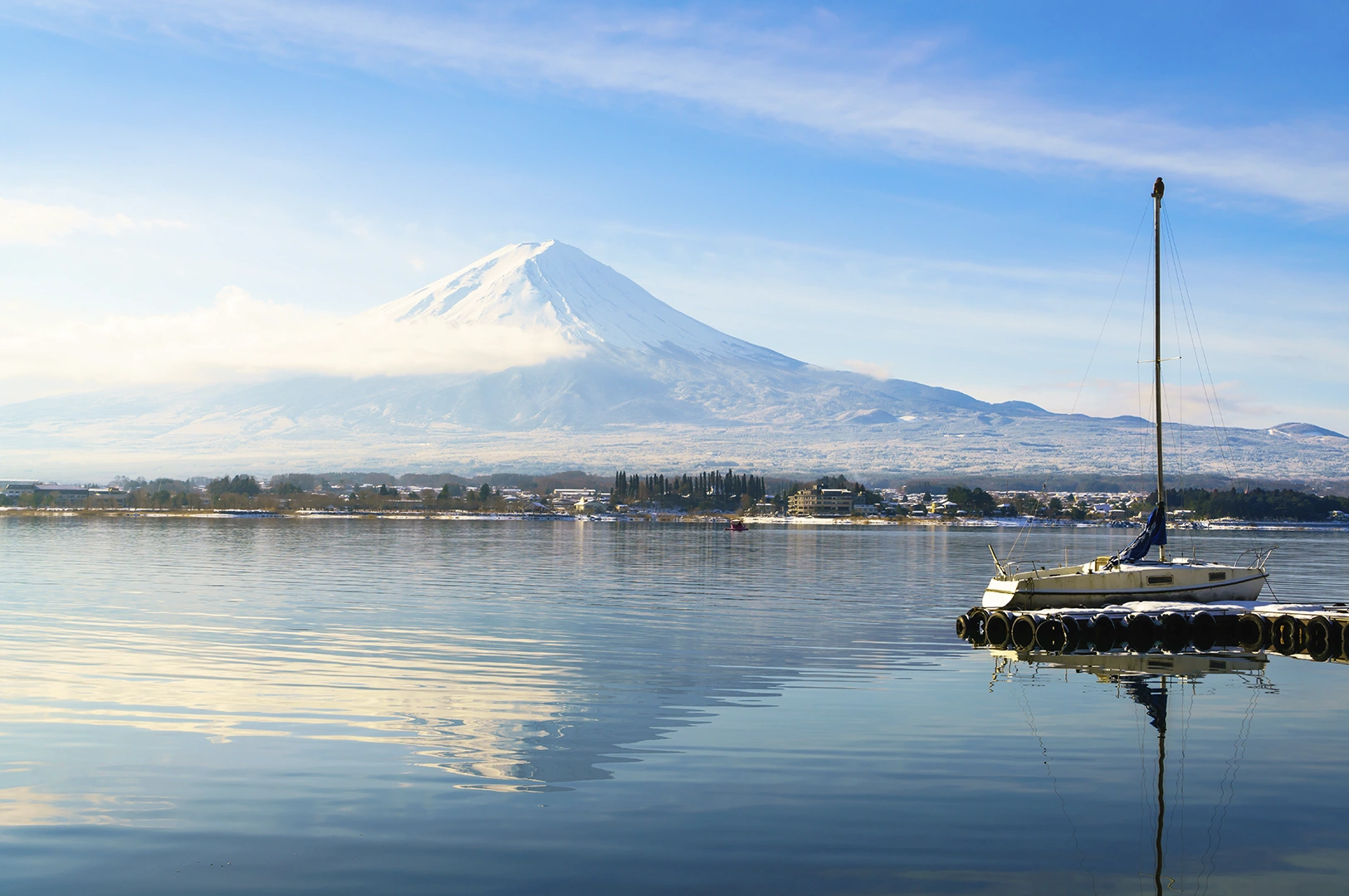 A serene, horizontal landscape of Mount Fuji, its snow-capped peak partially veiled by a thin horizontal cloud. The mountain is reflected perfectly in the mirror-like surface of Lake Kawaguchi. In the foreground to the right, a small white sailboat is moored at a dark, circular floating dock. The scene is bathed in soft, bright morning light with a pale blue sky and a light dusting of snow on the distant shoreline.
