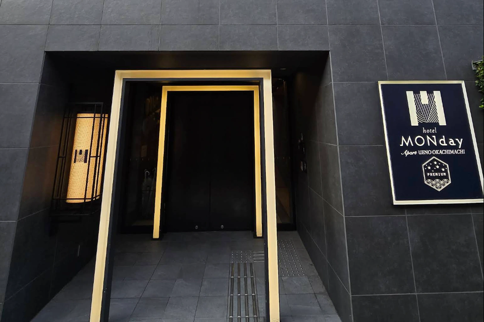 The sleek, dark grey stone entrance of Hotel MONday Apart Ueno Okachimachi. The doorway is framed by a minimalist, glowing yellow rectangular light box that creates a "portal" effect. To the right, a black vertical sign displays the hotel’s logo and name in elegant white and gold lettering.