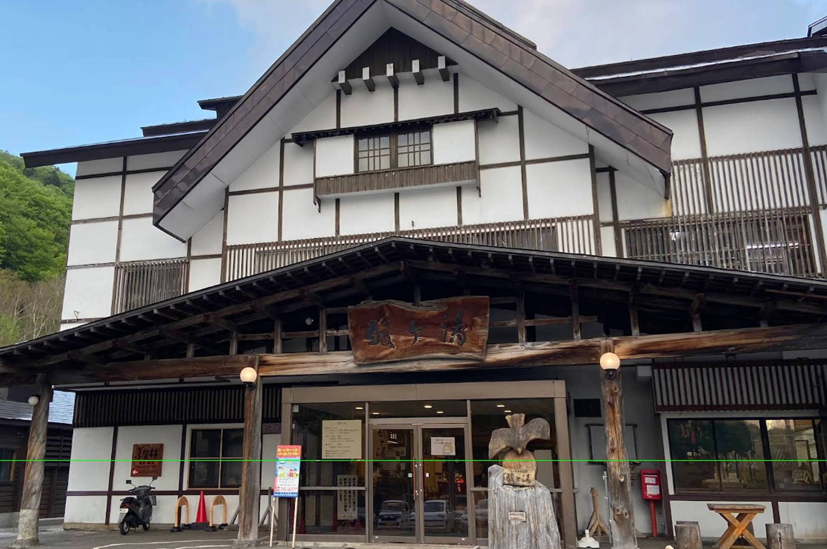The exterior of a traditional Japanese building with white walls, dark wooden beams, and a gabled roof. A sign with Japanese calligraphy is mounted above the main glass entrance. A wooden sculpture of a bird is positioned on a tree stump near the door, and a motorcycle is parked to the left.