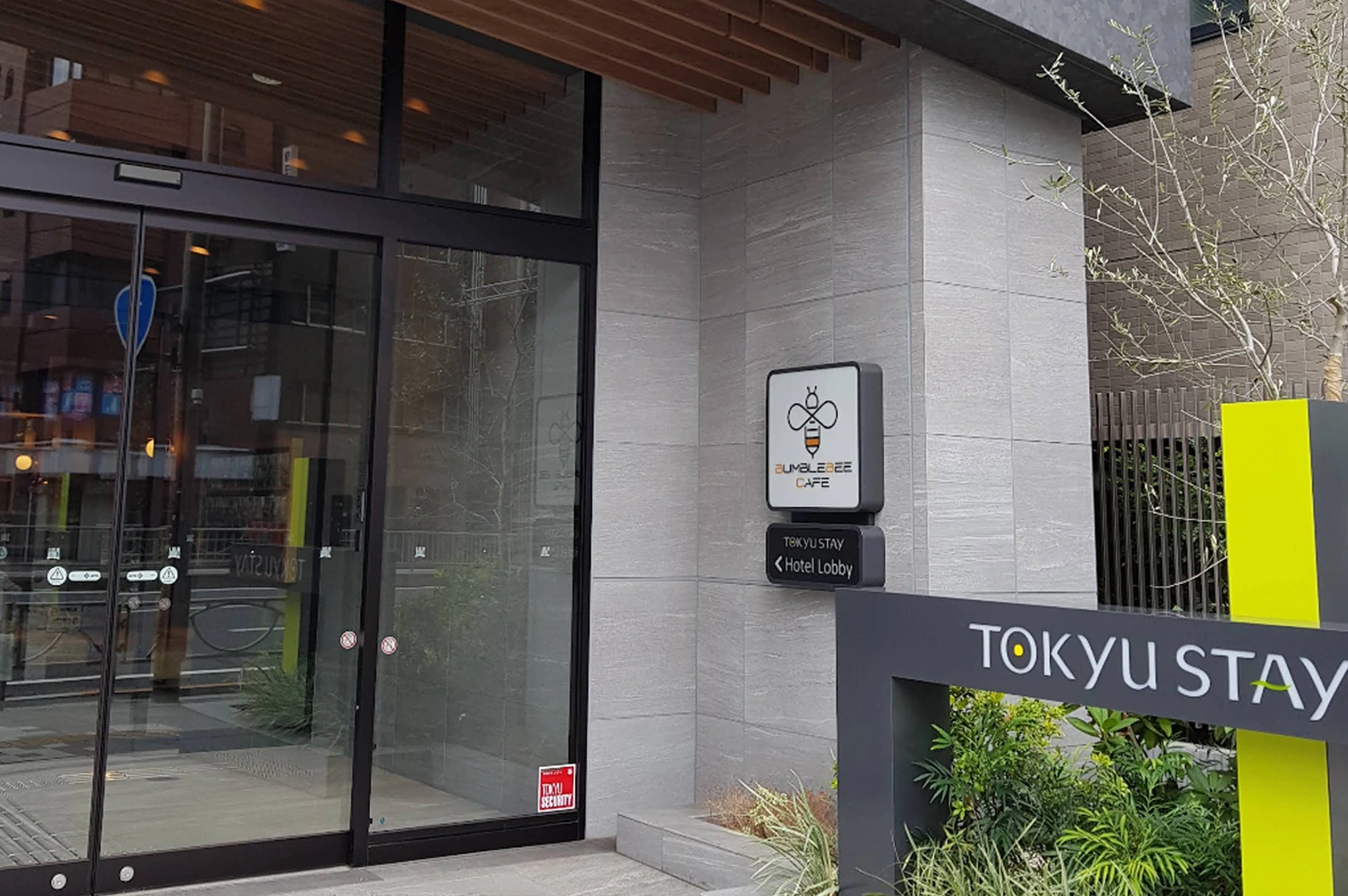 The exterior of a Tokyu Stay hotel highlighting the "Bumblebee Cafe." The building features light grey stone tiling and large glass windows. A sign for the cafe, featuring a minimalist bee icon, is mounted on the wall above a directional sign pointing to the hotel lobby. A bright lime-yellow accent pillar stands in the foreground by some greenery.
