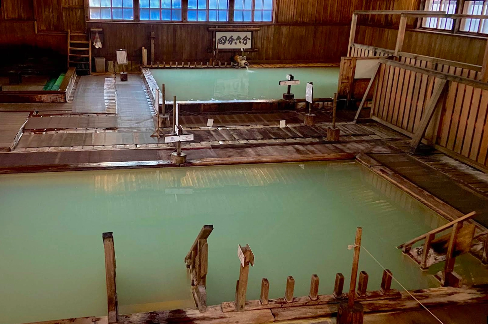 An indoor view of a traditional Japanese hot spring bath (onsen). Two large, rectangular stone pools are filled with opaque, light-green mineral water. The room features dark wood paneling, walls, and ceiling, with steps leading down into the baths, creating a rustic and relaxing environment.