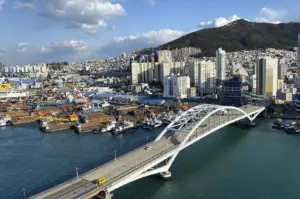 A high-angle, wide-set landscape showing the Yeongdodaegyo Bridge, a white arched drawbridge, stretching across a deep blue harbor. In the background, the city of Busan climbs up a steep, green mountainside with a mix of industrial shipyards, colorful cranes, and densely packed white and yellow residential high-rises. The sky is bright blue with scattered, fluffy white clouds, and a single yellow van is visible driving across the bridge.
