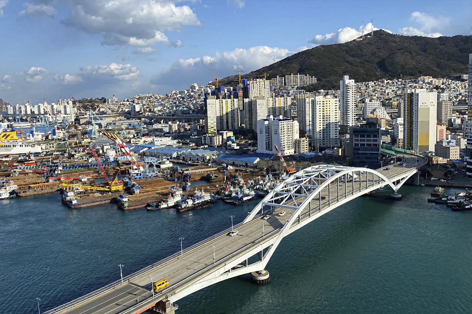 A high-angle, wide-set landscape showing the Yeongdodaegyo Bridge, a white arched drawbridge, stretching across a deep blue harbor. In the background, the city of Busan climbs up a steep, green mountainside with a mix of industrial shipyards, colorful cranes, and densely packed white and yellow residential high-rises. The sky is bright blue with scattered, fluffy white clouds, and a single yellow van is visible driving across the bridge.