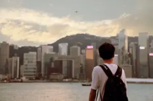 A contemplative, rear-view shot of a young man wearing a white t-shirt and a dark backpack, standing at the edge of a body of water and looking out toward a dense city skyline. The urban landscape, reminiscent of Hong Kong's Victoria Harbour, features a mix of towering skyscrapers and modern glass buildings set against a large, hazy hill in the background. The scene is bathed in the soft, warm glow of late afternoon or early evening light, with a pale, cloudy sky overhead where a small airplane is visible in the distance. The man’s centered position and still posture evoke a sense of reflection and the quiet transition of returning home after a period of travel.